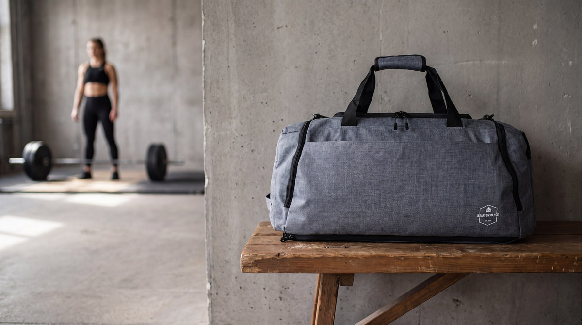 Graue Bearformance Ultimate Sportbag auf einer Holzbank vor einer Betonwand im Gym, im Hintergrund eine Sportlerin an der Langhantel
