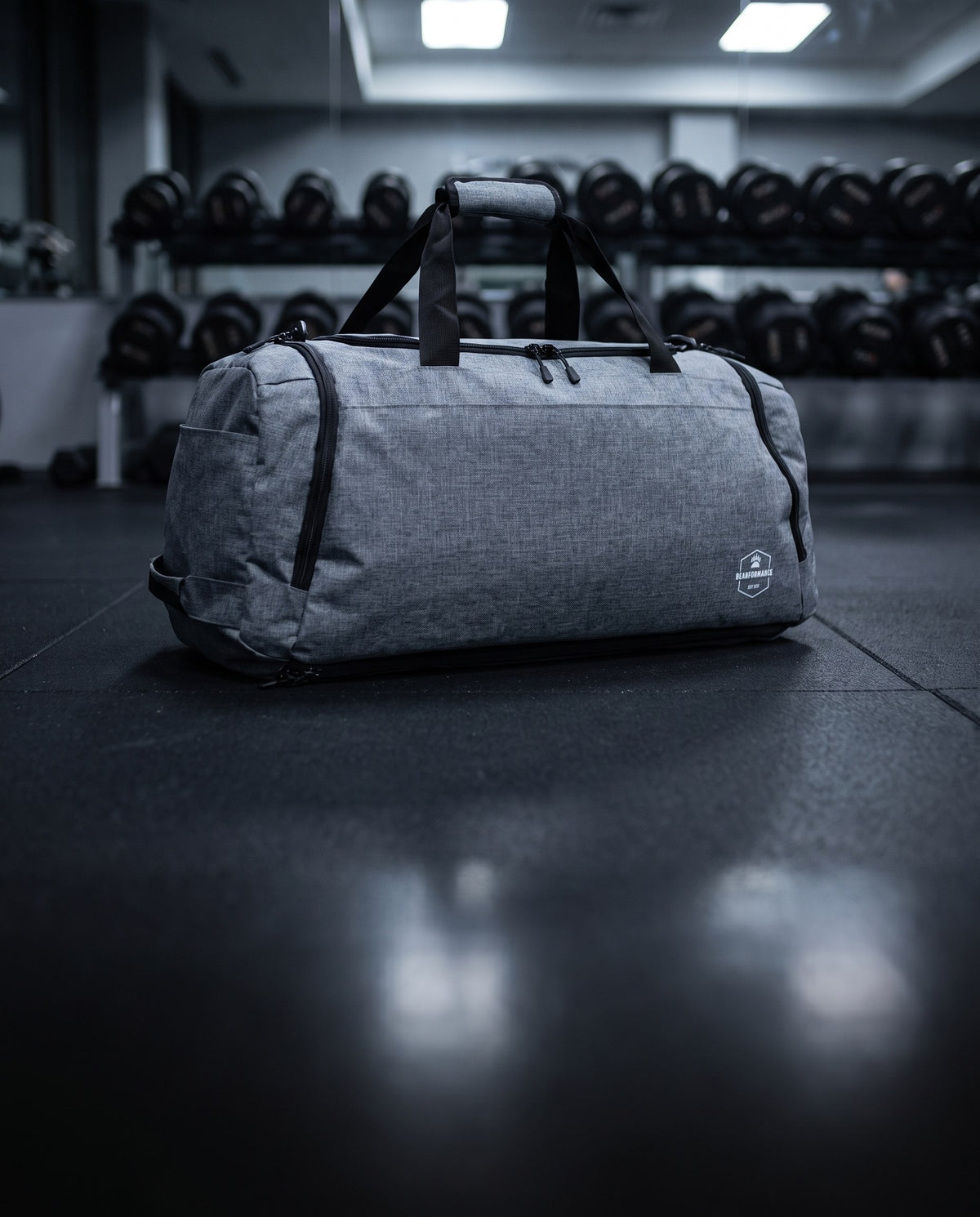 Graue Bearformance Ultimate Sportbag steht auf dem dunklen Boden eines Fitnessstudios, im Hintergrund ein Hantelregal